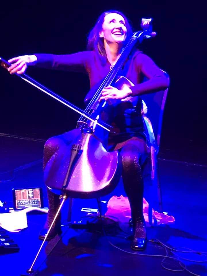 Jo-anne, seated playing her electric cello. The background is dark with deep purple overtones.