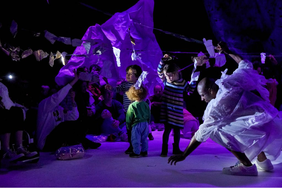 All Wrapped Up production image. Young audience members, joined by a performer, stick scraps of paper to strings of sellotape hanging overhead. The image is dark with purple tones.
