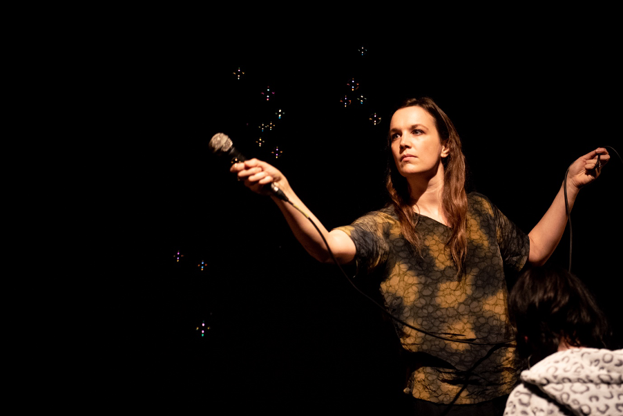 Rhiannon, a white person with long brown hair, is standing holding a microphone up to record the sounds of some bubbles popping.