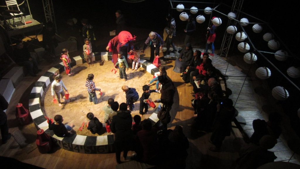 Aerial view of a performance of Mr and Mrs Moon. Performers and audience member play, make sandcastles and draw pictures together inside a big circular sand pit.