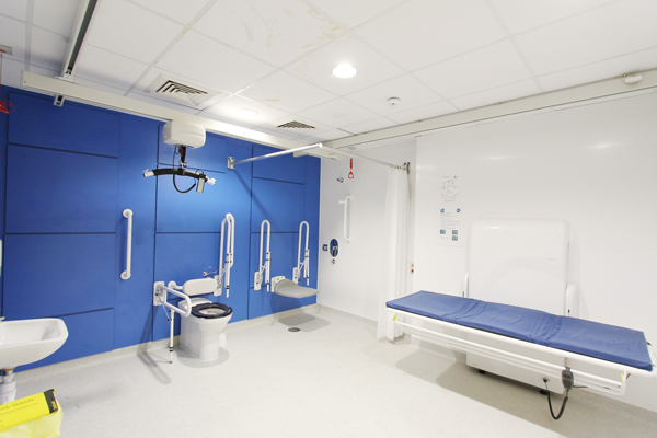 A spacious Changing places toilet. The far wall is blue, the wall to the right is white. There is a sink and bin on the left. A toilet and seat are on the back wall with grab rails and support walls. There is a hoist in the ceiling, and a blue change bench on the right.