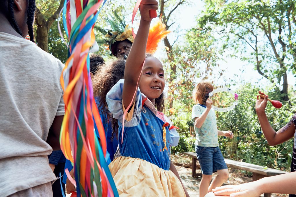 Child audience member hold string of colourful ribbons in the air. Other audience members holding percussion instruments surround them. Trees are behind them.