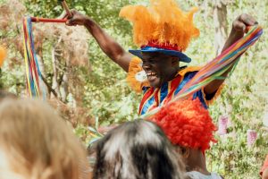 Smiling performer wearing colourful costume and a hat with big orange feathers. He holds a string of coloured ribbons between his outstretched arms. In the foreground are the heads of the audience. In the background are trees.