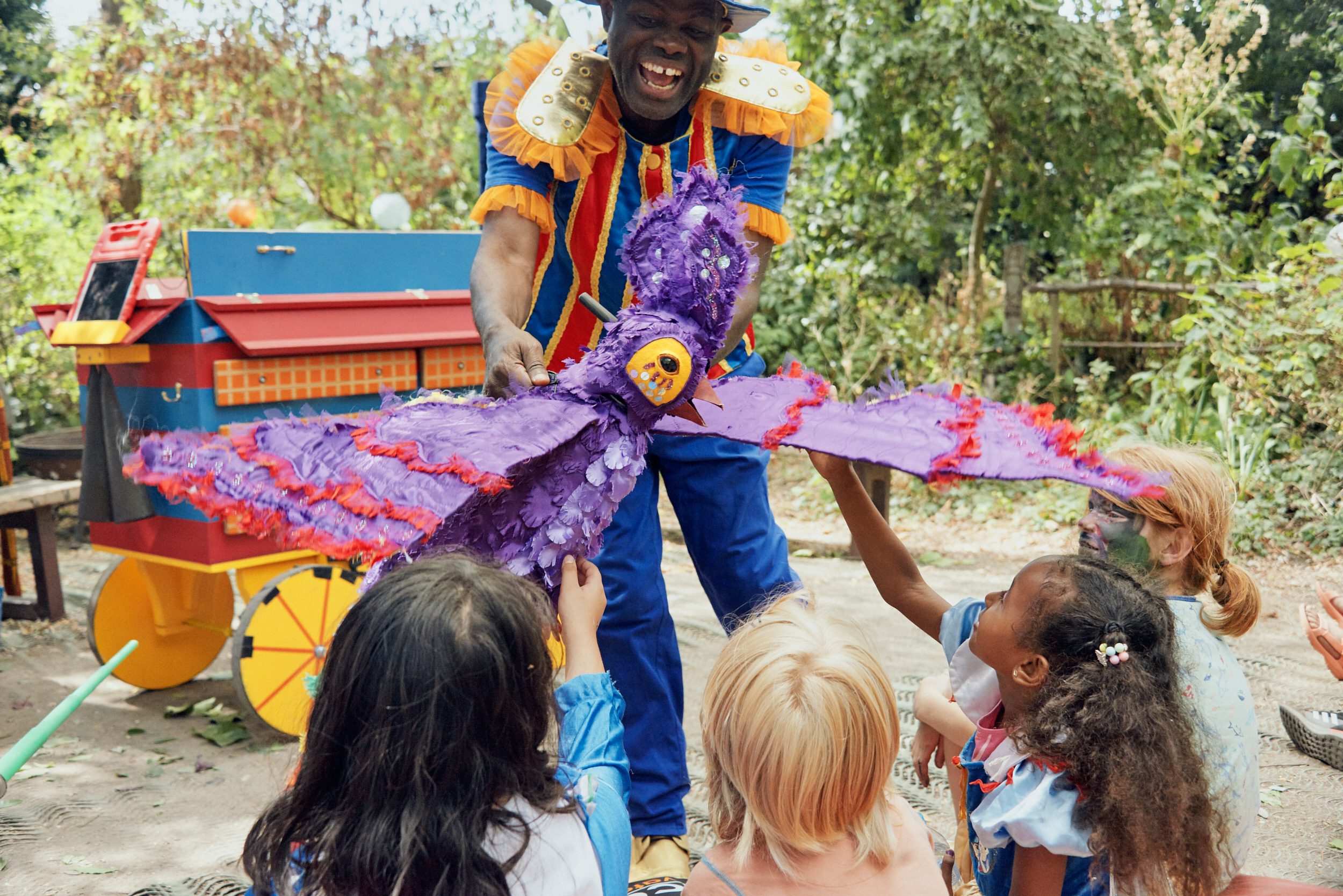 Smiling performer in brightly coloured costume holds a purple parrot puppet with outstretched wings over the heads of children in the audience. The children raise their arms to feel the parrot's wings. Behind the performer is a blue and red wooden cart with yellow wheels. The performance is outside, with trees all around.