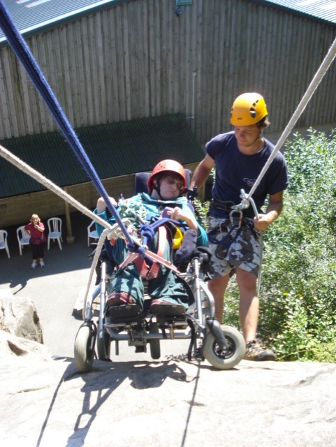 This photo is taken from the top of a climbing wall on a sunny day. It shows Mary in her late teens, wearing green trousers, a teal jumper and a red climbing helmet. There are ropes attached to her wheelchair with several knots and slings. Next to Mary is an instructor wearing shorts, tshirt and an orange climbing helmet. He has one hand on the handle of Mary’s wheelchair and one hand on the rope. Both Mary and the instructor look focused but relaxed. 