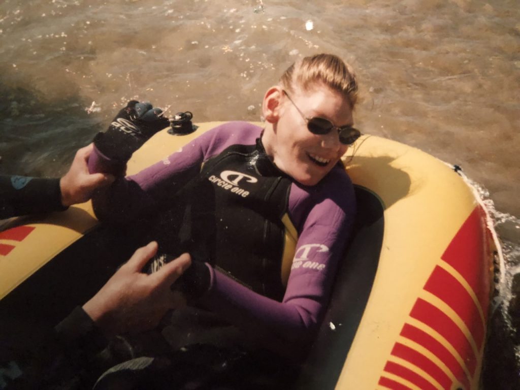 This shows Mary in her late teens, she has light brown hair pulled back into a ponytail and is wearing sunglasses. Mary is lying in a yellow and red dinghy,she is wearing a purple and black wetsuit and neoprene gloves cover her hands. Someone out of shot is holding Mary’s hands and both of their hands are in the picture. There is movement in the water around her and Mary has a broad smile, you can see her teeth and the dimples in her cheeks. 