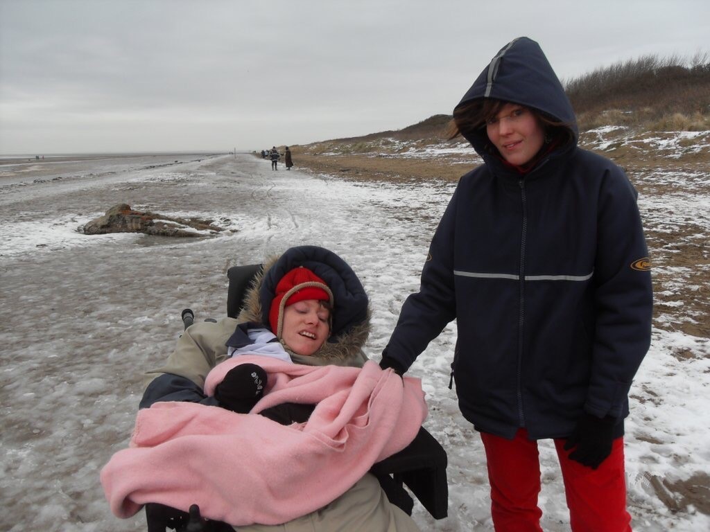 Two white women in their 20’s on a beach. The beach is covered in slushy snow. Behind the women you can see wiggly wheelchair tracks. One woman is wrapped up in a coat, with the furry hood up and a red woolly hat underneath, she has a big pink blanket on her lap and is sitting in a wheelchair, this is Mary. The woman next to her is standing up, they are holding hands. This woman is also wrapped in a thick coat with the hood up. This is Esther. They look cold but they are smiling.