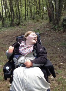 Mary is pictured here in her early 30s in some woodland in early Spring. Mary is seated in an electric wheelchair, with a pale leg cosy covering her lower half. Mary is looking upwards and to her left with a beaming smile on her face. 