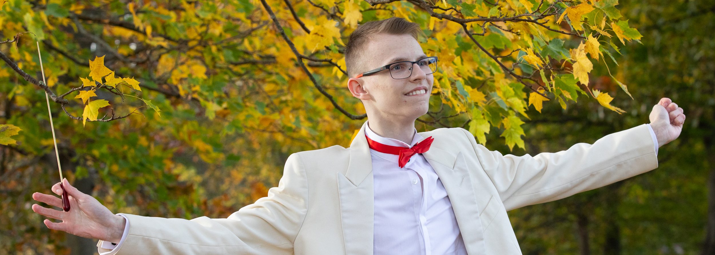Teenage boy stands outside. He wears a white coat and tails with a red bow tie. He holds his arms wide, with a conductor's baton in his left hand. Behind him are trees with yellow autumnal leaves.