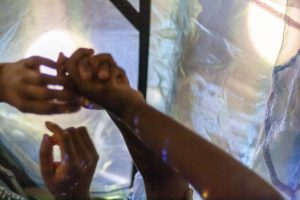 Adult and child arms raised in the air, holding hands. Behind the hands is green-blue gauze material with light shining through it.