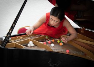 Woman in a red dress sits under the opened lid of a grand piano. She plays with colourful ping pong balls on the piano strings.