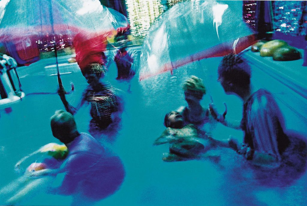 A blurry, stylised photo of performers holding see-through umbrellas over aucience members in a hydrotherapy pool. Though the muted blue tones of the photo make it difficult to see defined items, there are flashes of colour from the performer's costumes and the instruments on the edge of the pool.