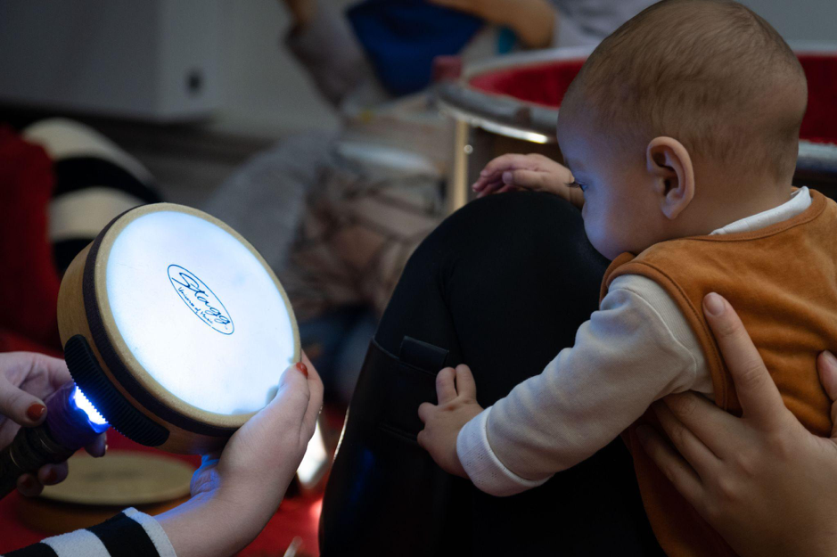 A baby held leaning on a parent's knee looks at a drum with torch light being shone through it.