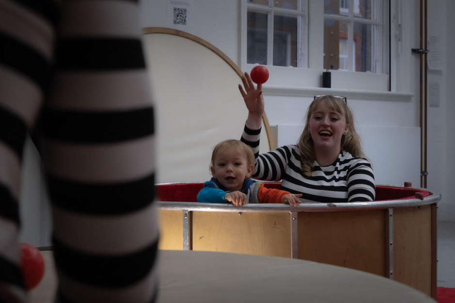 Siobhan, a blond white woman in her 20s wearing a black and white striped top, sits inside a drum-shaped ball pool. Next to her is a baby holding onto the edge of the ball pool. Siobhan is throwing a ball out, towards a figure in the foreground also wearing black and white.