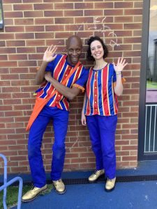 A photo of two people in red, blue and gold striped tops, blue trousers and gold shows. On the left is a Black man with a shaved head, with his right hand in the air waving. On the right is a white woman with short dark brown hair with her left hand in the air waving. Both are smiling. They are standing in front of a brick wall.