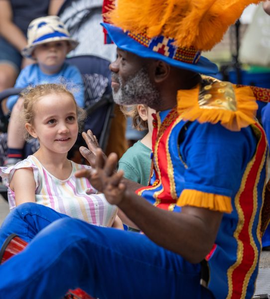 Young audience sit alongside storyteller Usifu, looking up and listening to him. Usifu is in a bright blue and orange costume, gesticulating with his hands as he tells the story of The Lost Feather.