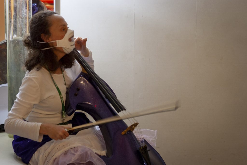 Jo-anne is relaxed and sitting with her purple sparkling cello, wearing a white top, purple sparkly material around my waist and a full white skirt overlapping a netted vintage skirt. Jo-anne's brown shoulder length hair is pulled back and she is wearing a see-through face mask. She is bouncing her bow and looking towards something happening in the room (not pictured)