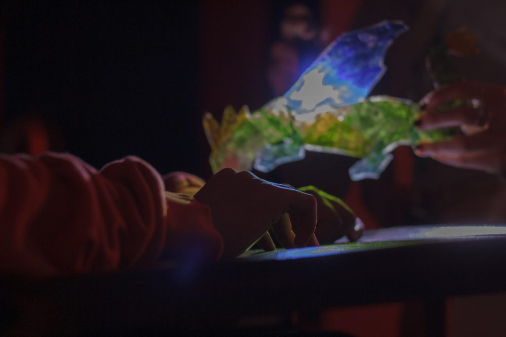 This is a close up shot, the room is dark, a young person's hands are resting on a tray table, and hovering just above the tray table is a translucent green blue and yellow dragon puppet. A supporter holds the puppet with one hand and shines a light on its wings, illuminating the dragon and bathing the young person's hands and the tray table in colour.