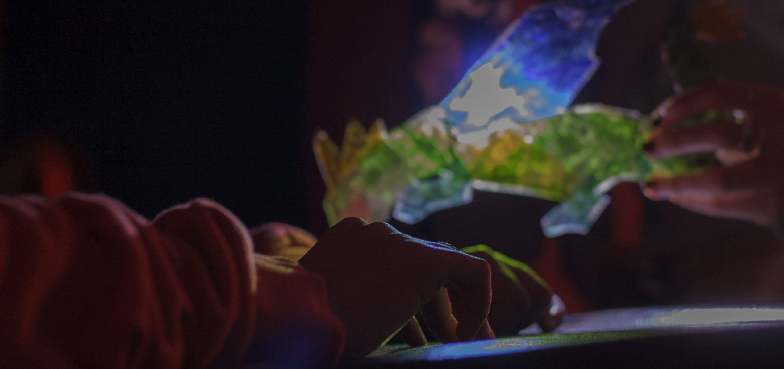 Young person’s hands rest on tabletop top. Beyond them, a light shines through a colourful dragon shadow puppet.