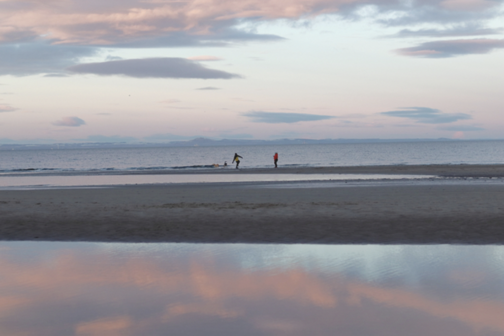 Two people play with their dogs on a sandy beach under a light pinky blue cloudy sky at sunset. There are reflections of the clouds and pink sky in a watery pool on the beach. It's cold out so the people wear bright jackets. One is throwing something into the sea.