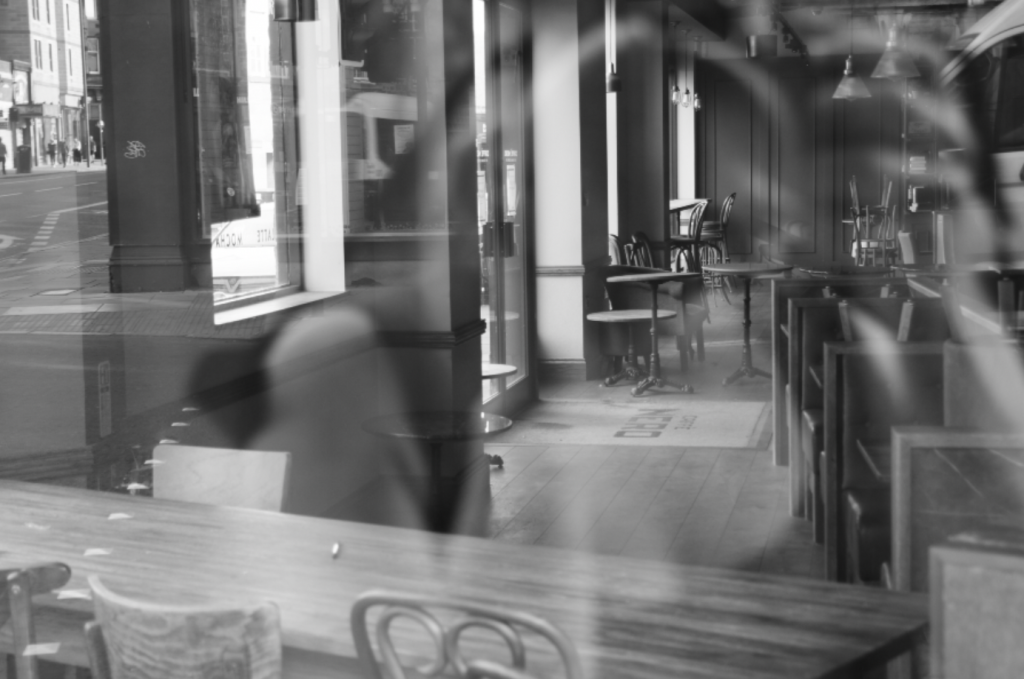 Image description: An empty bar or cafe during the first lockdown. The photograph is taken through the window and with the photographer’s reflection in the pane of glass. In focus is an empty wooden chair next to a wooden table facing another window opposite. There are chairs stacked upside down on other tables and empty armchairs. Through the window on the opposite side of the cafe is an empty street with people walking in the distance. The photo is in black and white.
