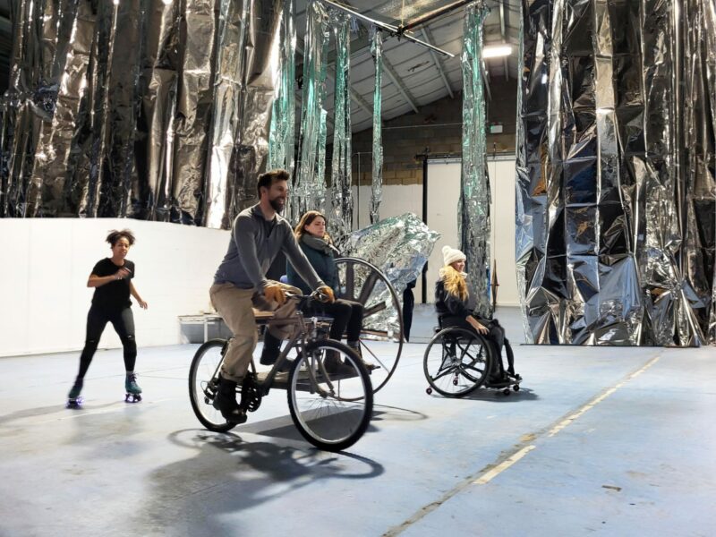 Two members of the creative team riding on a mock-up fantastical vehicle. A performer using a wheelchair and a performer on roller skates ride alongside and behind them. They are in a large rehearsal space. There is silver material in sheets and strips hanging down from above, a mock up of a possible performance installation space.