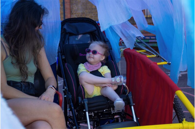 An adult and young child, seated in a mock up of a fantastical performance vehicles. They are gazing happily at each other. Blue material is draped from above, encircling the vehicle.