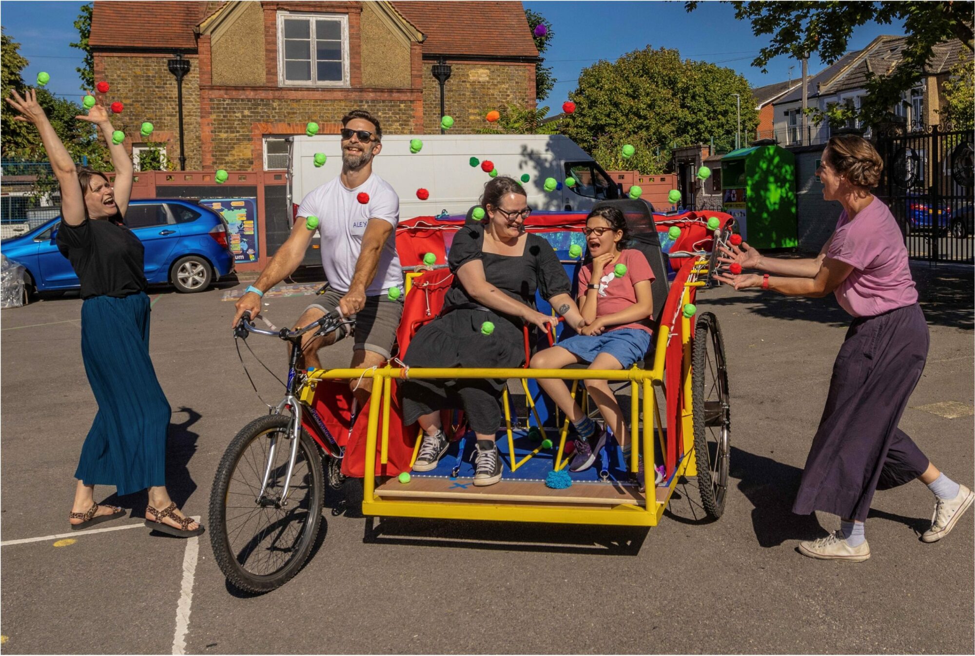 A photo from the rehearsal and development of Joyride. An adult and young person are seated next to each other in a specially-created vehicle. They are outside in a school playground. The vehicle has a yellow bar across the front, and a folded-down red canvas roof and big wheels at the back. On the right, the vehicle is attached to a bicycle being ridden by a man in a white shirt and sunglasses. Two women stand on each side of the vehicle as it passes. Their arms are raised jubilantly as they release colourful balls into the air.