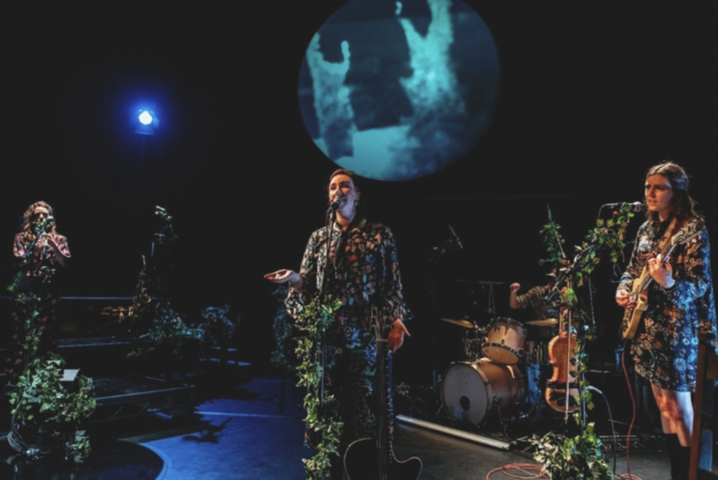 A four-piece band performing on a dimly lit stage. Claire is in the centre, singing. Each performer is wearing an outfit using the same patterned material of flowers and plants. The mics and other bits of staging are wrapped in ivy.