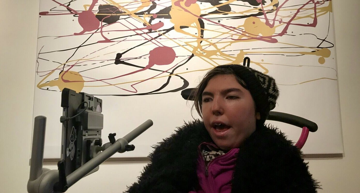In this photo Greta is sitting in her wheelchair in front of one of her paintings hanging on the wall. She is a young white woman with brown hair and she is wearing a patterned beanie hat with a pink jumper and fluffy black jacket. The painting behind her is a mix of yellow, pink and black swirls and lines on a white background.]