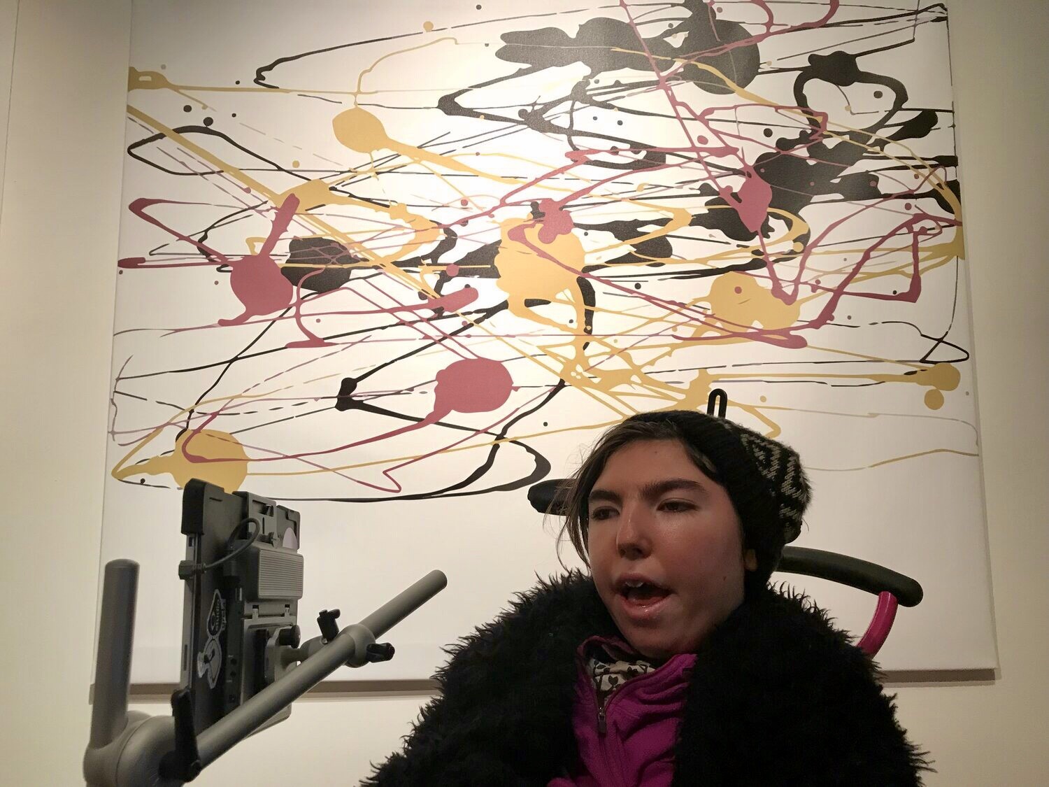 In this photo Greta is sitting in her wheelchair in front of one of her paintings hanging on the wall. She is a young white woman with brown hair and she is wearing a patterned beanie hat with a pink jumper and fluffy black jacket. The painting behind her is a mix of yellow, pink and black swirls and lines on a white background.]