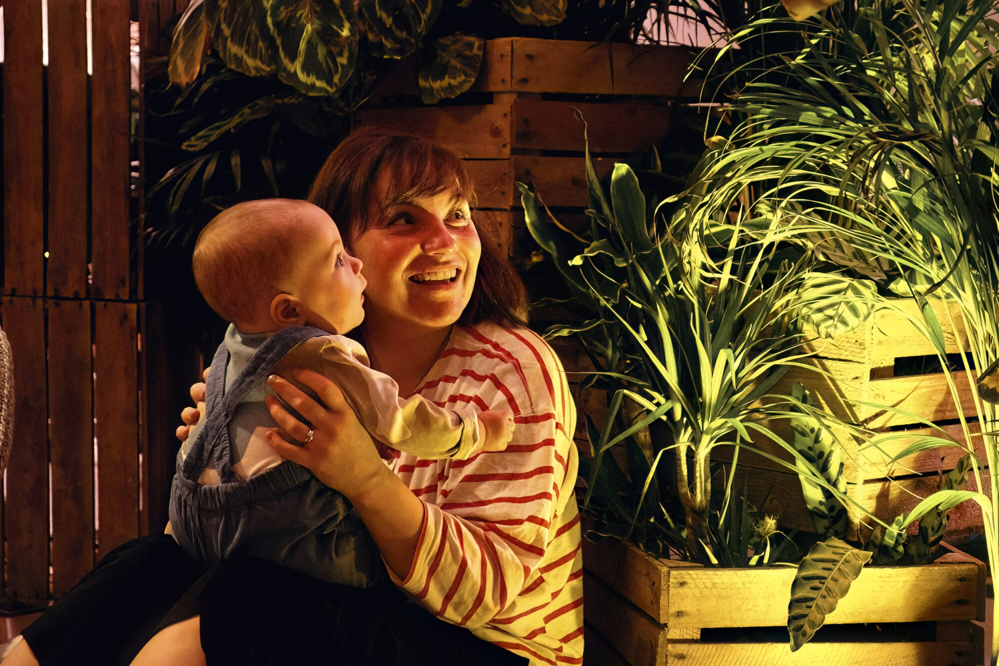 A smiling mother holds a baby facing her on her knees. They are both looking with wonder across at something out of shot. Behind them are crates filled with plants. The lighting illuminates their face with a golden glow.