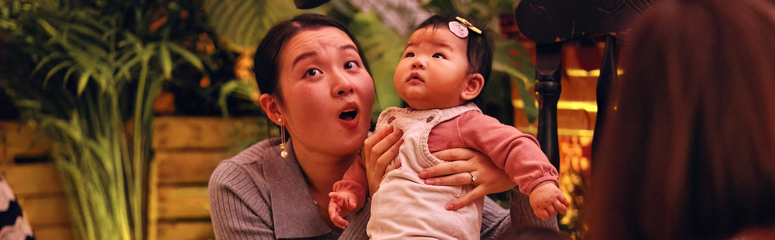 A mother sits on the floor, supporting a baby to sit on a chair next to her. They are both looking up at a speaker above them with wonder. Behind them are wooden crates filled with plants.