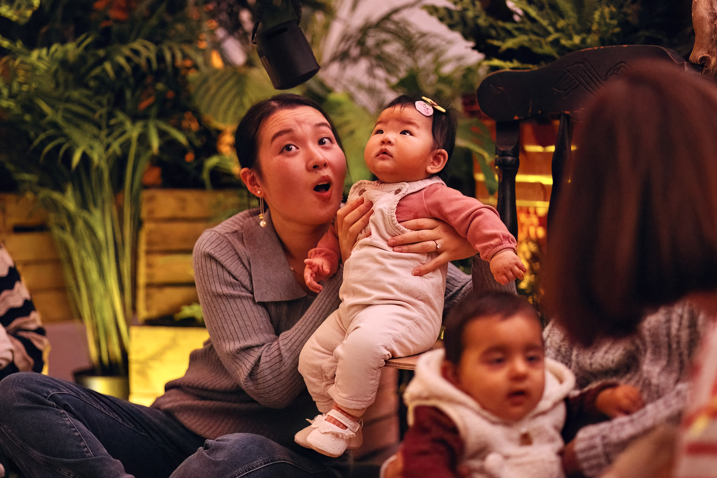 A mother sits on the floor, supporting a baby to sit on a chair next to her. They are both looking up at a speaker above them with wonder. Behind them are wooden crates filled with plants.