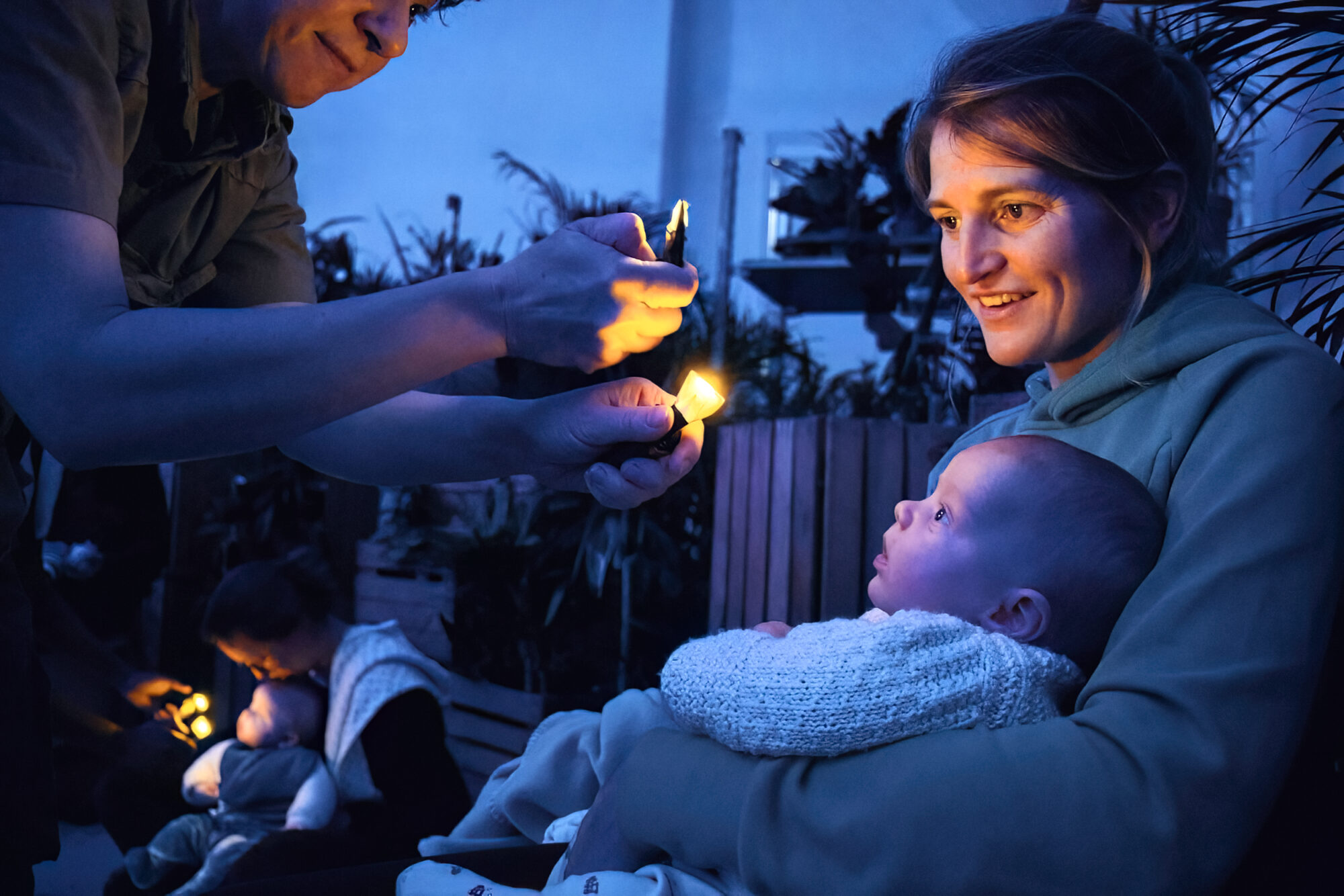 A mother holds a baby in her arms. They both look up in wonder at small firely lights held towards them by a performer. In the room's blue light, the mother and baby's faces are illuminated by the lights.