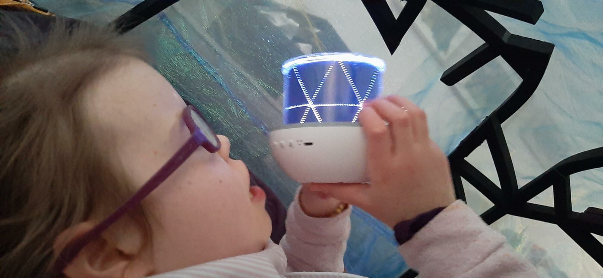 Lucy is a white girl with purple glasses and brown hair tied back. She is holding up a bespoke nightlight to cast shadows on the blue gauzy tent material overhead. This is part of At Home show, Space to Be.