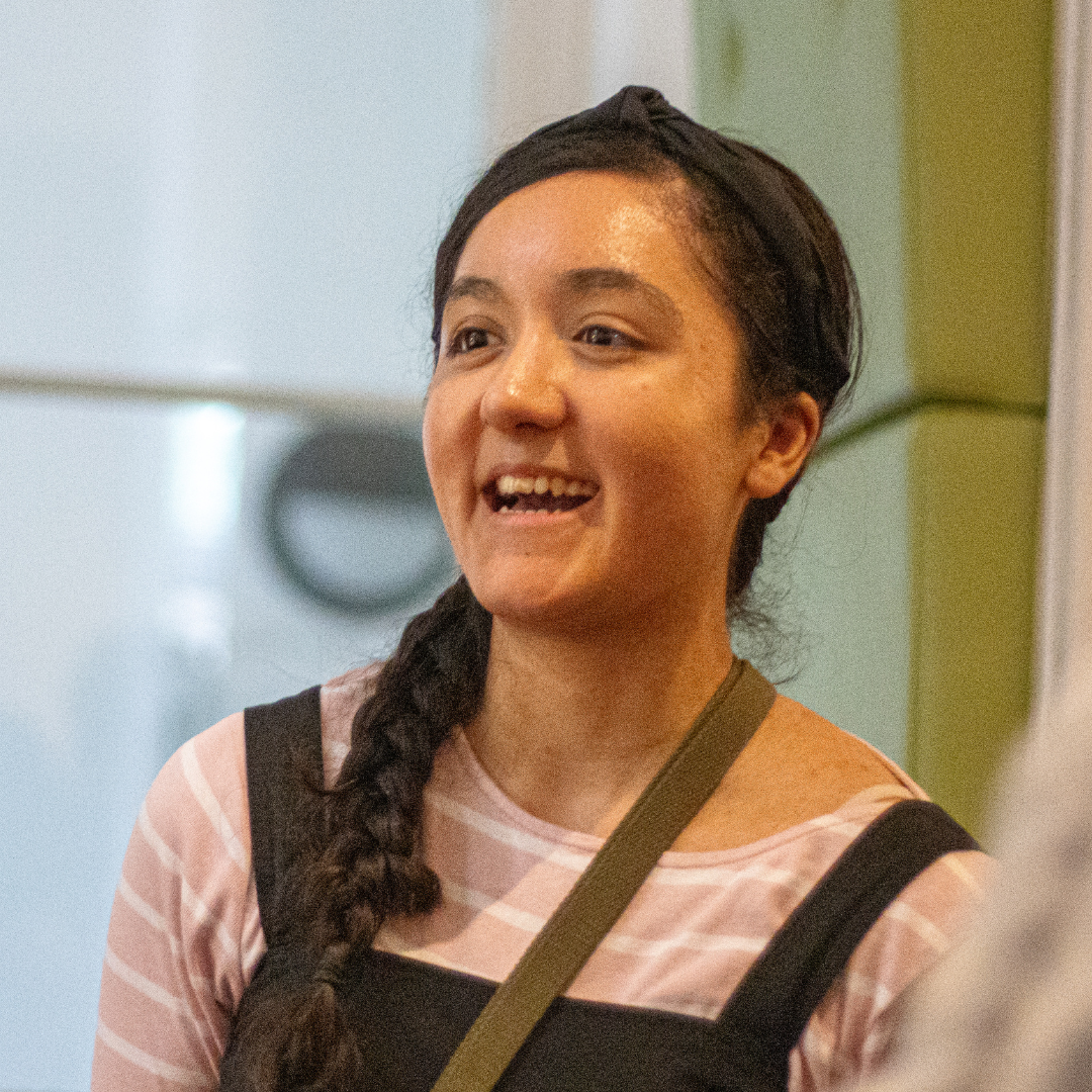 A photograph of Shania, a mixed raced Sri Lankan woman in her early-mid 20's. She has long black hair in a plait along her right shoulder. She is wearing a black headband, a strip pink-white long sleeve top, black dungarees and a small green bag with the strap across the left shoulder. In the photo, she is smiling and happy, looking as though she is having an engaging conversation with another fellow peer. The image was taken at a networking event run by peer-led group, Young Arts Fundraisers.