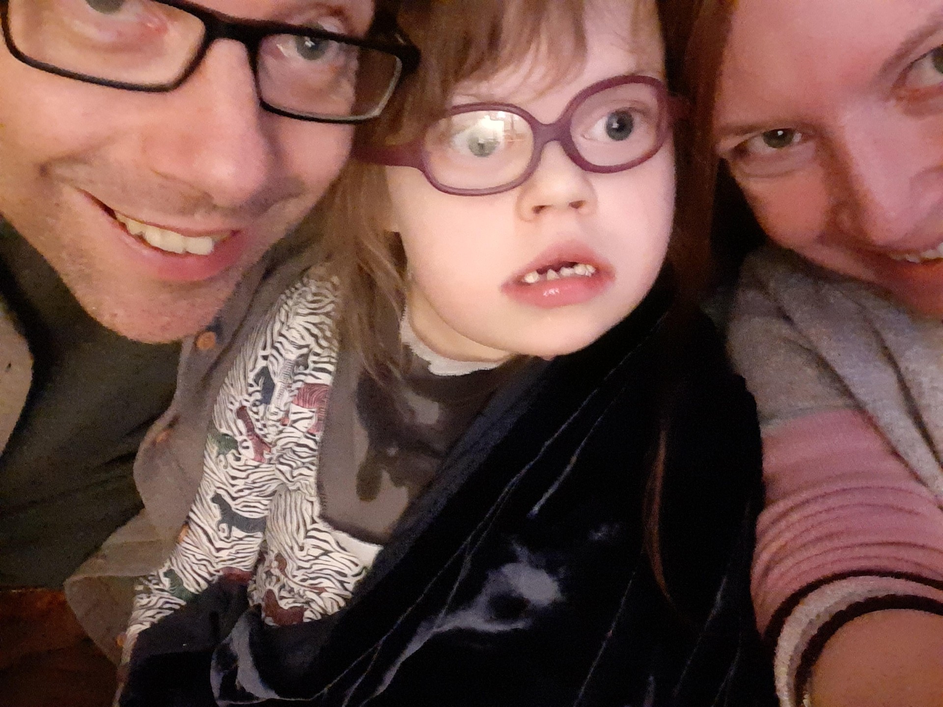 Sam with her partner and daughter Lucy. It's a selfie, with their heads close together looking into the camera. Lucy has a sensory blanket from At Home show Space to Be wrapped around her shoulders.