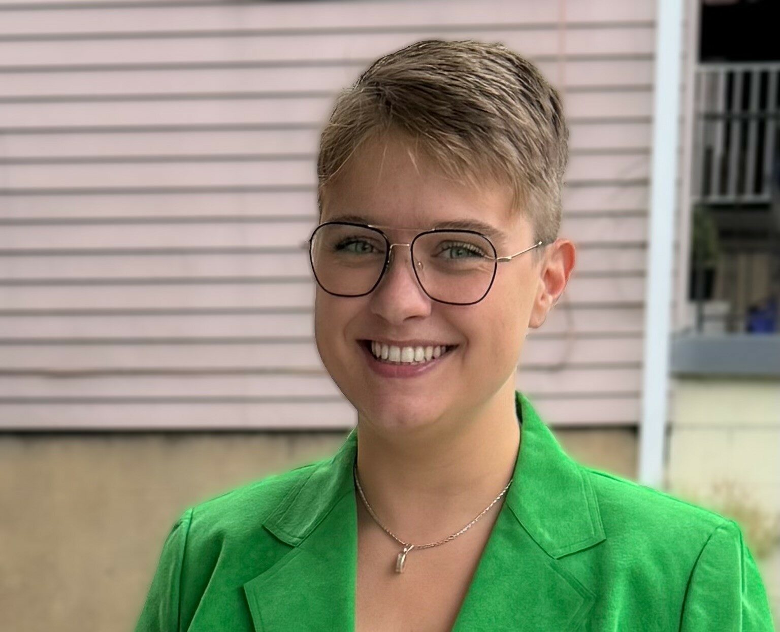 Alison Mahoney (she/they) has blond cropped hair, wire glasses and a green blazer. They smile at the camera. Behind them a brown building with clapperboard details is visible.