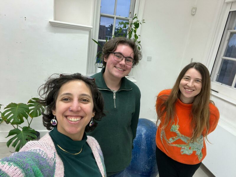 From left to right: Access and Wellbeing Officer, Maka; Placement Student Billie; and Executive Director, Zoë. They smile widely. They are standing in the rehearsal room at Oily Cart, a white studio space with tall windows, plants and a speckled large blue musical egg prop behind them.