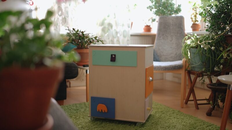 A chest of drawers, except the drawers are all different colours and shapes, and are on different sides. The chest is in the middle of a living room, which is filled with plants.