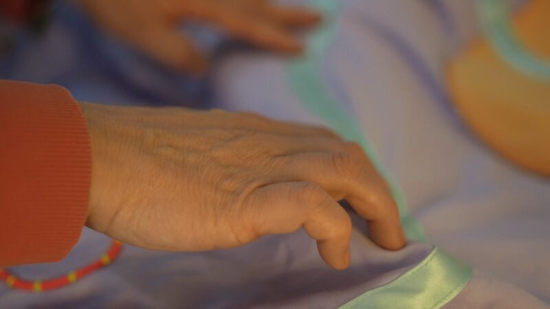 Lilac material, with shapes and patterns made of different colours and material sewn into it. Fingers feel along a blue ribbon running down the middle.Fingers following a long blue ribbon, sewn onto lilac material.