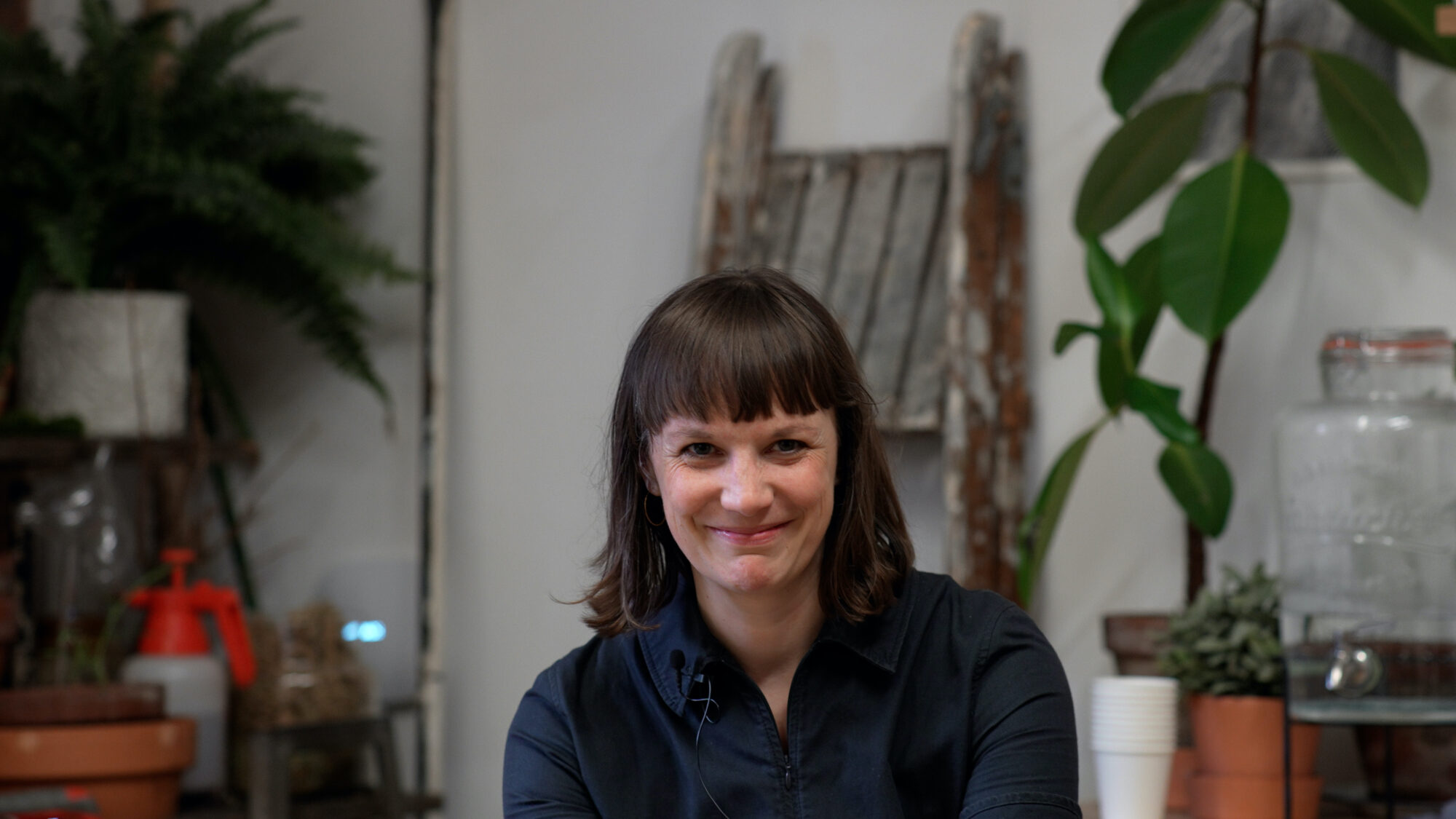 Rhiannon is a white person with a brown bob and blunt fringe, wearing a black shirt. They are smiling and looking intently into the camera. They are a in a room - maybe an art studio - with white walls, a paint covered wooden ladder leant against a wall, and various plants and pots.