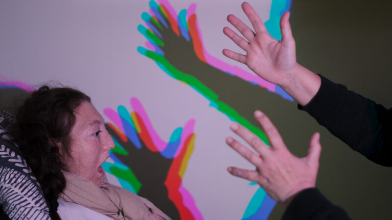 A young person looks art an adult's hands which are raised to make shadows against the wall. A special bulb means the shadow has colourful, overlapping outlines in purple, green, blue and pink