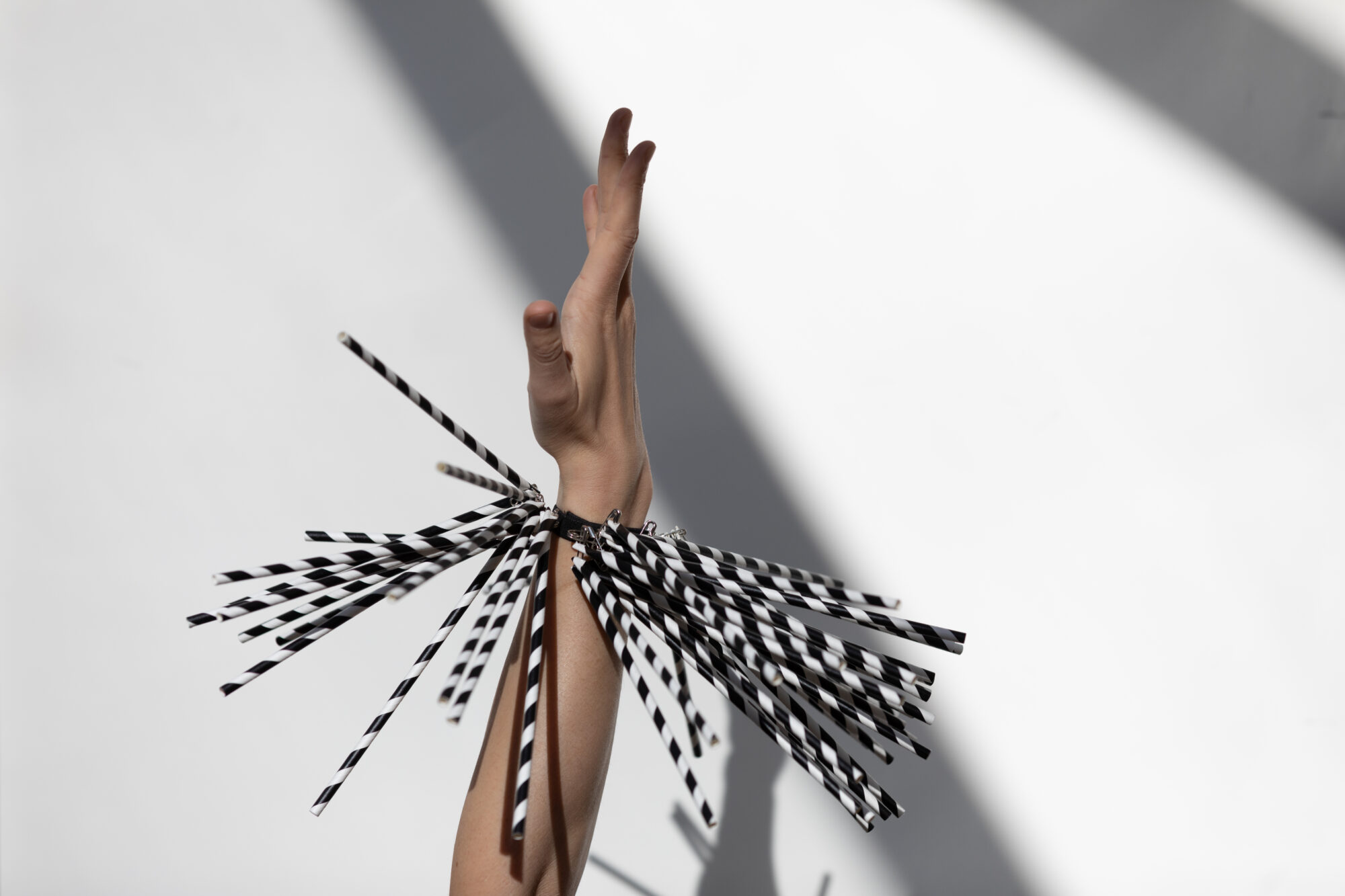 An extended white arm with fingers outstretched. On the wrist is a bracelet of black and white straws. There is a sense of movement, as though the arm has just twisted, sending the straws to wave outwards in different directions. The diagonal black and white stripes of the straw are mirrored by the white wall in the background, with diagonal stripes of shadow.