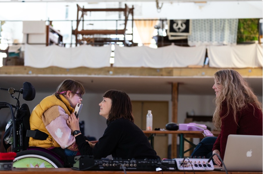 Rhiannon, a white adult with a brown bob and blunt fringe, kneels in front of session musician Lucy, a white teenager with pink glasses, a yellow gilet and using a wheelchair. Lucy and Rhiannon look intently at each other, as Rhiannon holds a tambourine to Lucy's mouth. Lucy's mum Sam sits close by. Sound equipment is in the foreground. 