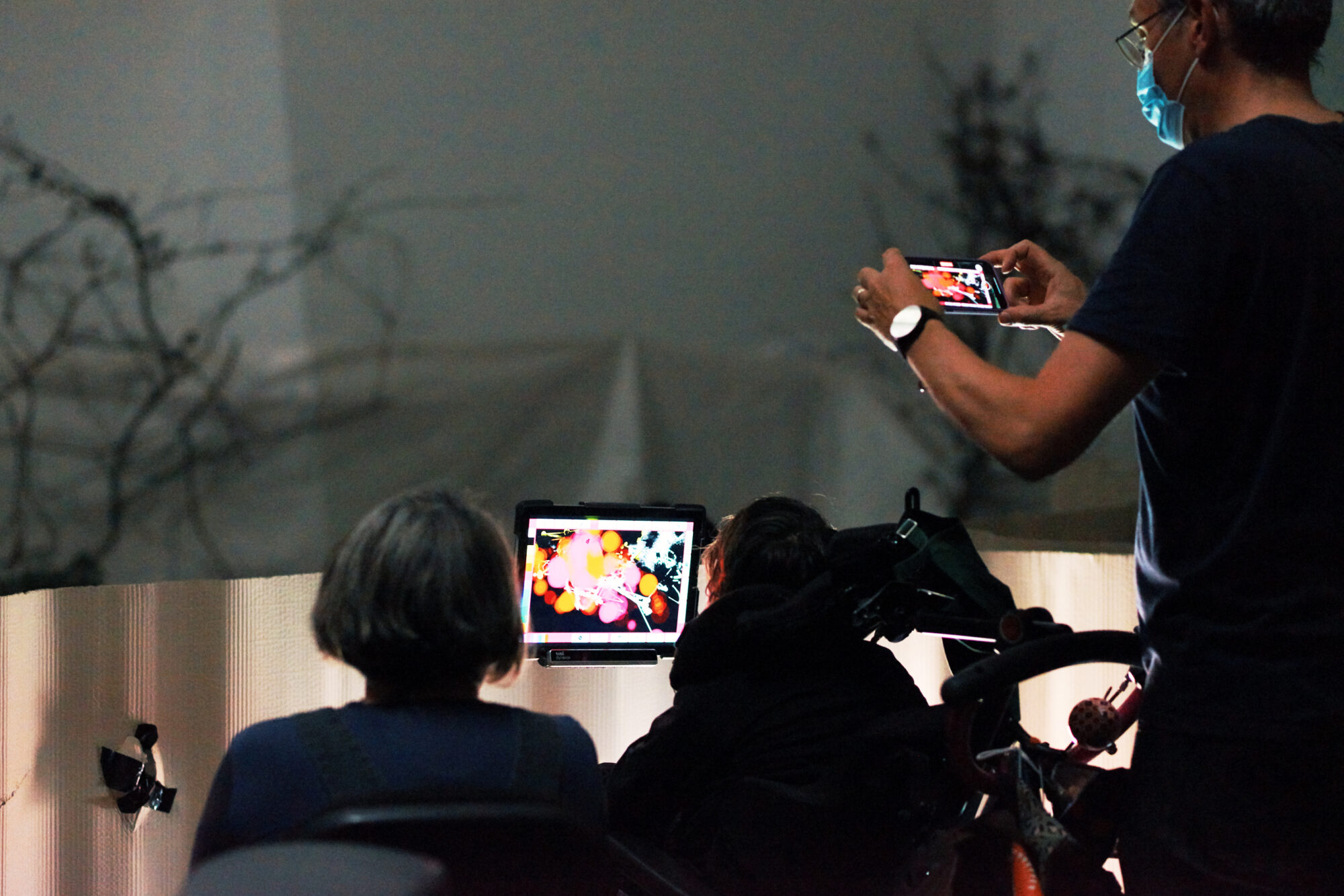 Young artist Greta creates an artwork of vibrant white, pink and orange, on the eyegaze screen that is attached to her wheelchair. Her mum sits next to her, and her dad stands above, taking a picture of the art. They are in a dimly lit room, with a corruagedt cardboard sheet surrounding them. In the distance, branches reach up the white walls.