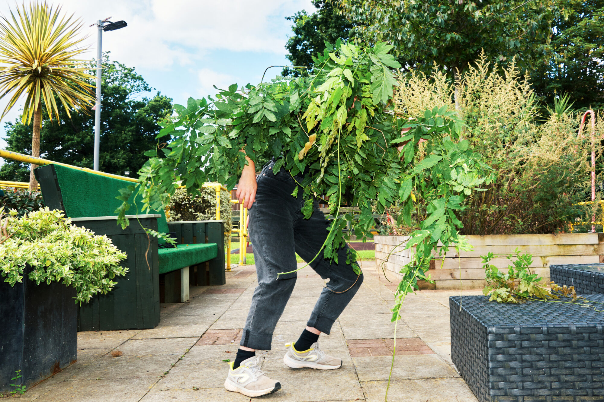 A person stands outside in a plant-filled patio surrounded by trees. The top half of their body is covered with leaves and branches, resembling the shape of a head, chest and arms. Their lower body and legs are visible, and bent as if they are about to run away.