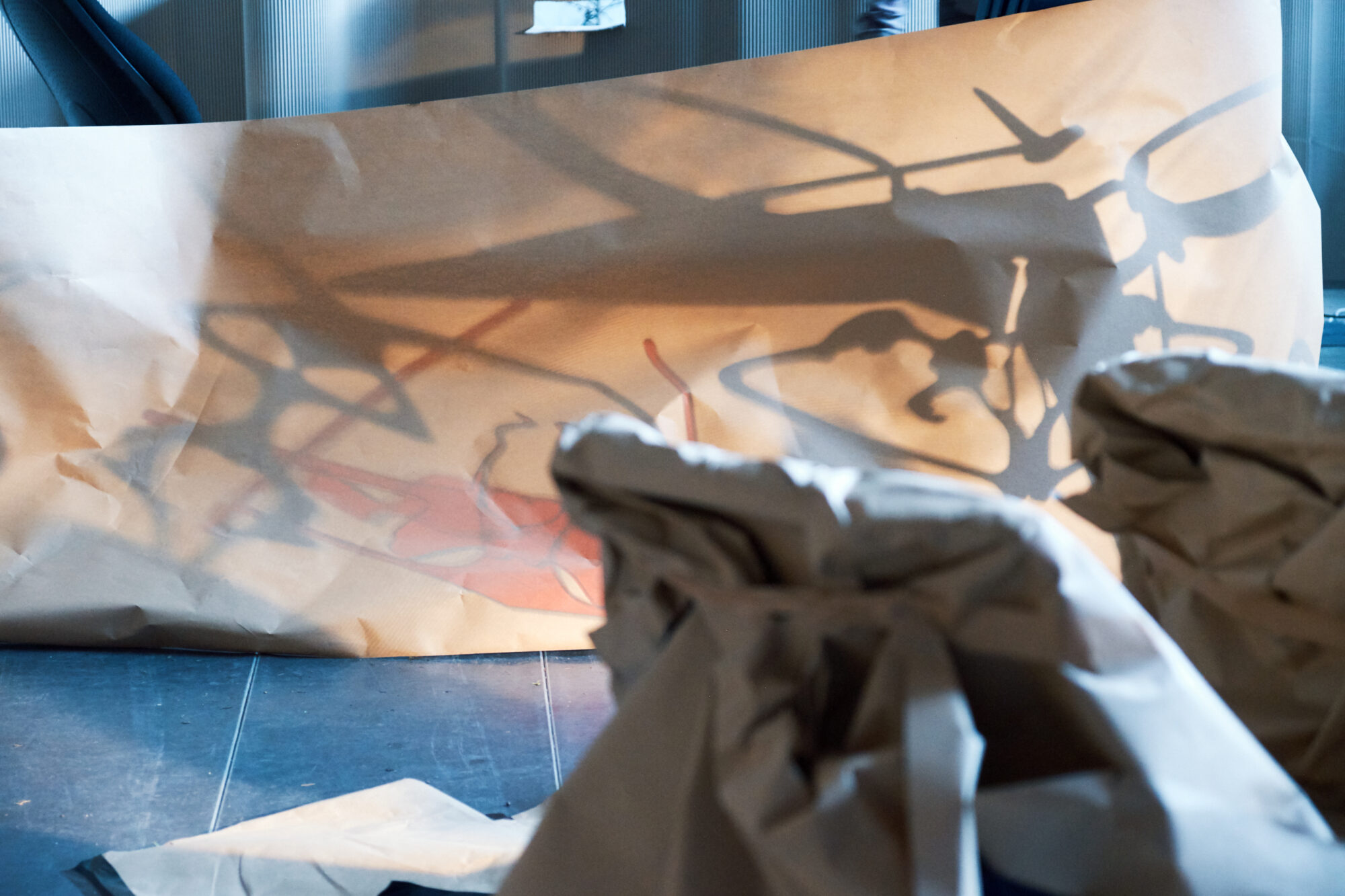 Red and black shadow shapes appear through a long stretch of brown paper. A ball of scrunched up paper is in the foreground.