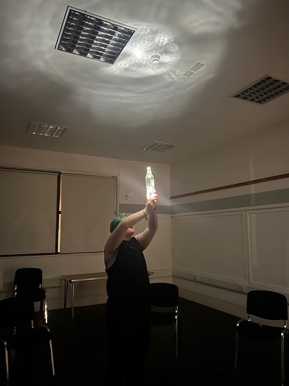 Billie holds up a clear glass bottle filled with water. An unseen light underneath the bottle magically casts rippling shadows onto the white ceiling above.